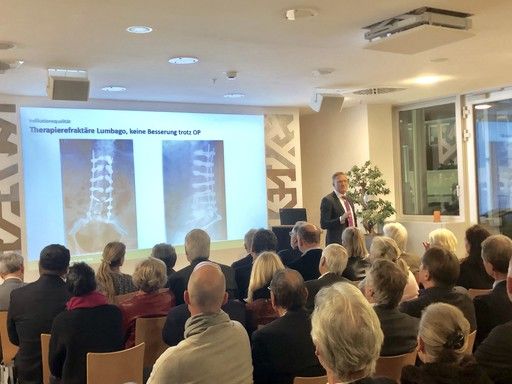 Ein Referent steht vor einem Publikum und präsentiert zwei Röntgenbilder, die eine Therapie bei therapieresistentem Lumbago zeigen. Der Text auf der Präsentationsfolie besagt: "Therapieresistente Lumbago, keine Besserung trotz OP." Das Publikum besteht aus zahlreichen Zuhörern, die aufmerksam zuhören.