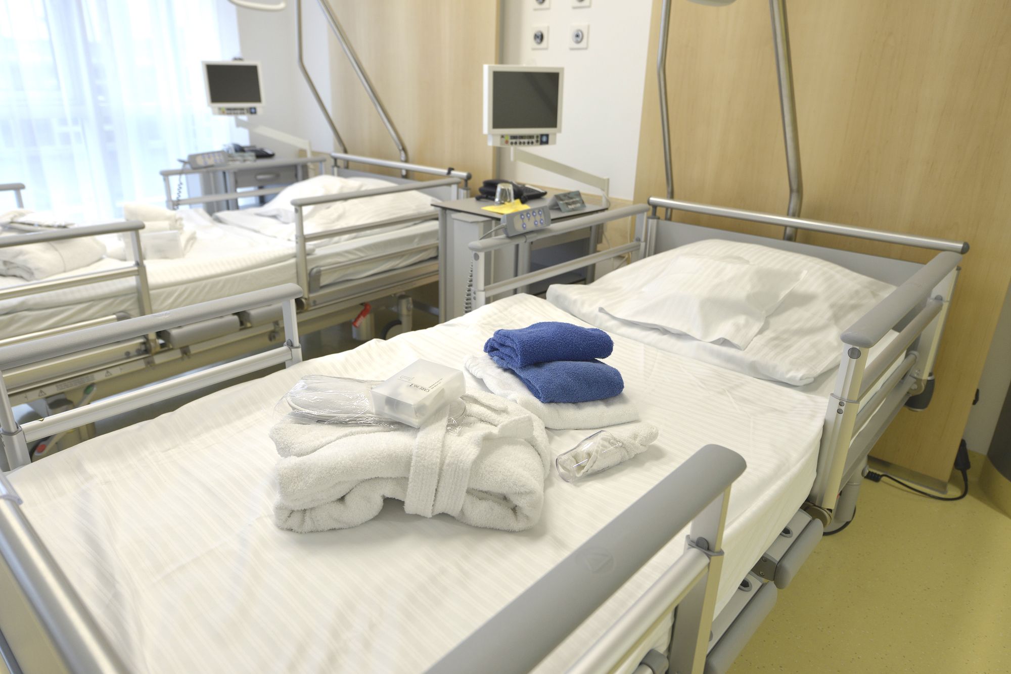 A hospital room features multiple beds, each with neatly arranged white linens. On one bed, folded towels are displayed: white towels along with a blue towel. Monitors and medical equipment can be seen in the background, indicating the setting's healthcare purpose.