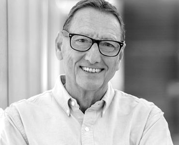 Ein älterer Mann mit Brille lächelt freundlich in die Kamera. Er trägt ein helles Hemd und hat kurzes, graues Haar. Der Hintergrund ist unscharf, wodurch der Fokus auf dem Gesicht des Mannes liegt.