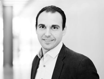 Das Bild zeigt einen Mann mit dunklen Haaren und einem freundlichen Lächeln, der in einem professionellen Outfit posiert. Der Hintergrund ist unscharf und hell, was den Fokus auf sein Gesicht lenkt.