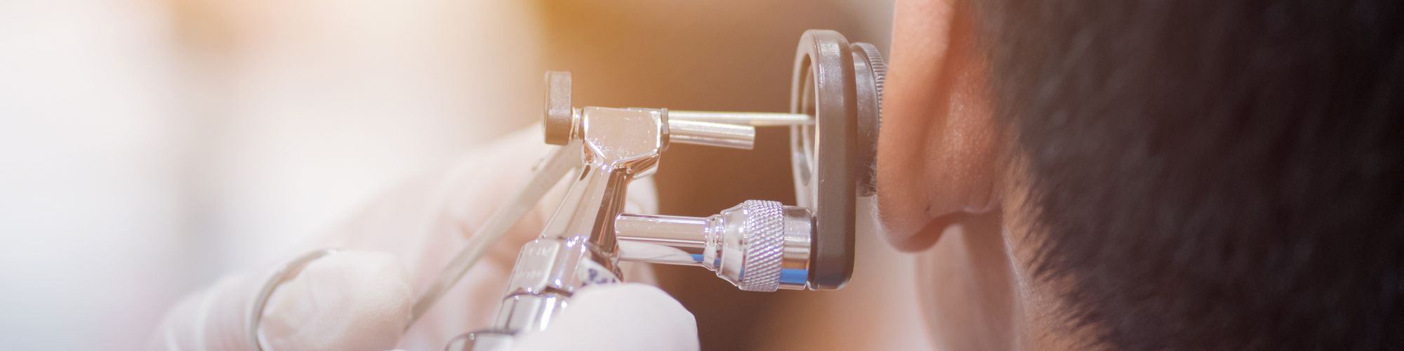 A close-up image shows a healthcare professional examining a patient's ear with an otoscope. The otoscope's light illuminates the ear canal, highlighting the medical procedure taking place. The professional's hands are gloved, indicating a sterile environment.