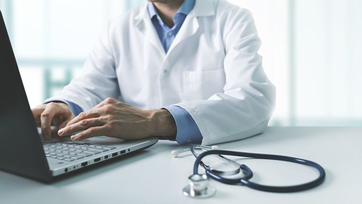 A healthcare professional in a white lab coat types on a laptop. A stethoscope rests on the table beside them. The background suggests a clinical or office environment.