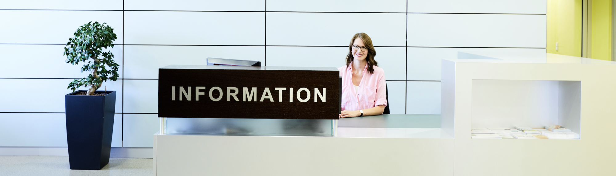 A receptionist sits at an information desk in a modern, well-lit space. The desk prominently displays the word "INFORMATION." A small potted plant is positioned beside the desk, contributing to a welcoming atmosphere.