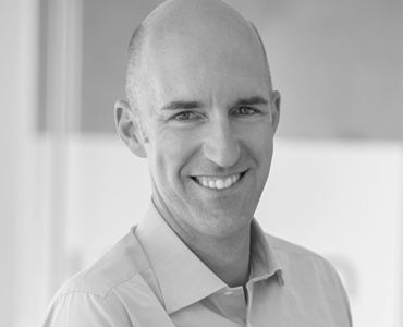Ein lächelnder Mann mit Glatze trägt ein helles Hemd. Er steht vor einem hellen Hintergrund, der unscharf ist. Sein Gesichtsausdruck vermittelt Freundlichkeit und Offenheit.