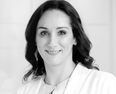 Das Bild zeigt eine lächelnde Frau mit langen, dunklen Haaren, die einen weißen Laborkittel trägt. Sie steht in einem hellen Umfeld und wirkt freundlich und professionell.