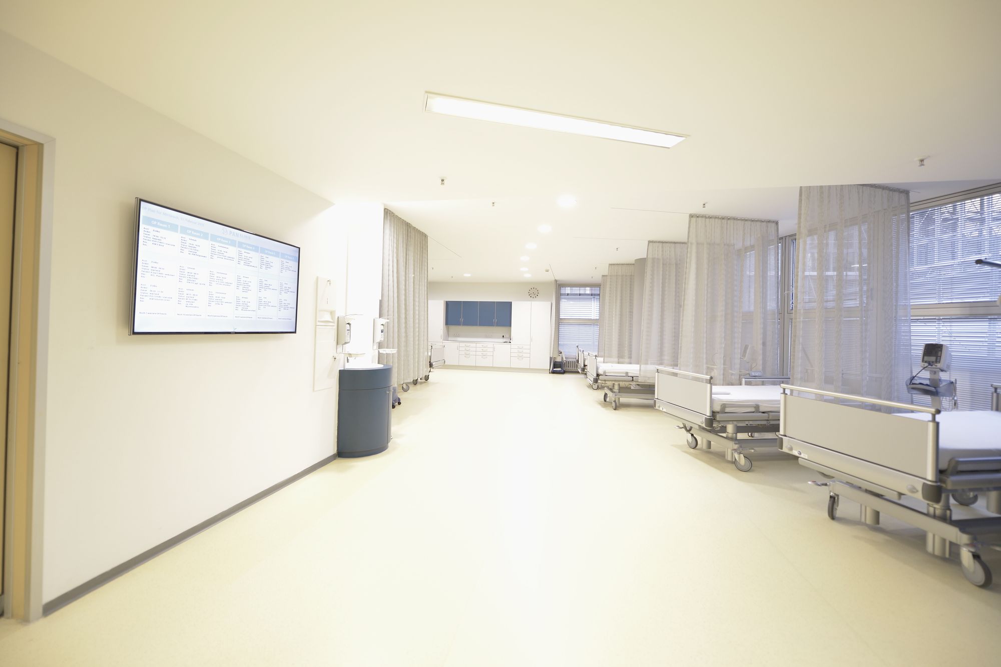 A brightly lit hospital corridor with white walls and a smooth floor. On one side, there are hospital beds on wheels, separated by curtains. A wall-mounted screen displays information, with a water dispenser nearby. The atmosphere is clean and clinical.