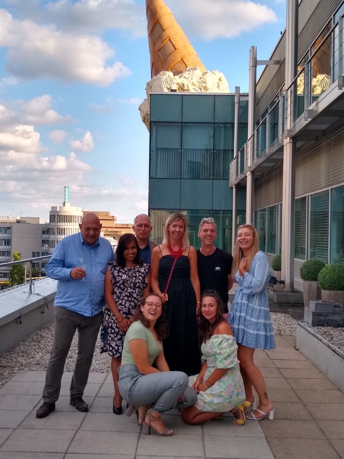 Eine Gruppe von sieben Personen steht lächelnd auf einer Terrasse. Im Hintergrund ist ein Gebäude mit großen Fenstern und einem auffälligen, riesigen Eistüten-Dekor zu sehen. Der Himmel ist blau mit einigen Wolken. Die Stimmung wirkt freundlich und entspannt.
