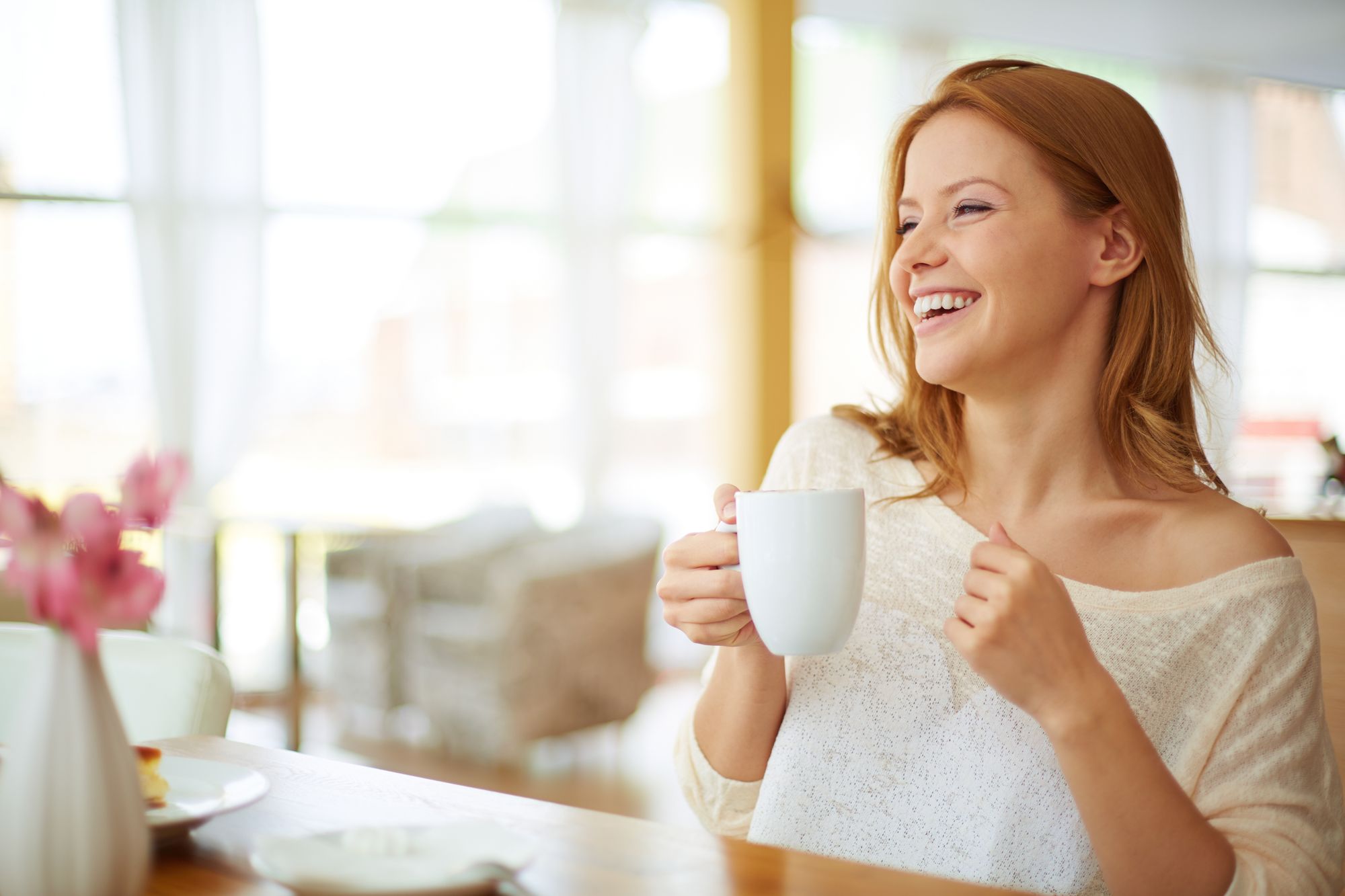 A smiling woman sits at a table holding a white coffee cup, enjoying a moment of warmth and relaxation. She wears a light sweater and appears cheerful, suggesting a comforting breakfast or coffee break. Soft natural light illuminates the background.