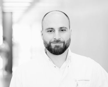 Ein Mann mit Bart trägt einen weißen Arztkittel. Er schaut direkt in die Kamera und wirkt freundlich. Der Hintergrund ist verschwommen und hell, was eine professionelle Atmosphäre vermittelt.