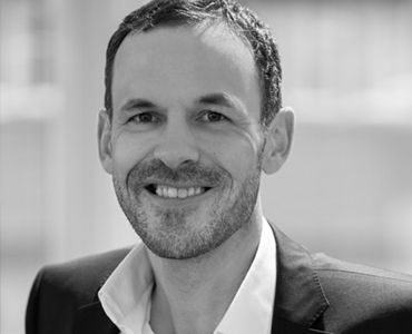 Ein lächelnder Mann mit kurzem Haar und einem gepflegten Bart trägt einen schwarzen Anzug mit einem weißen Hemd. Der Hintergrund ist unscharf und hell. Die Gesichtszüge wirken freundlich und offen.