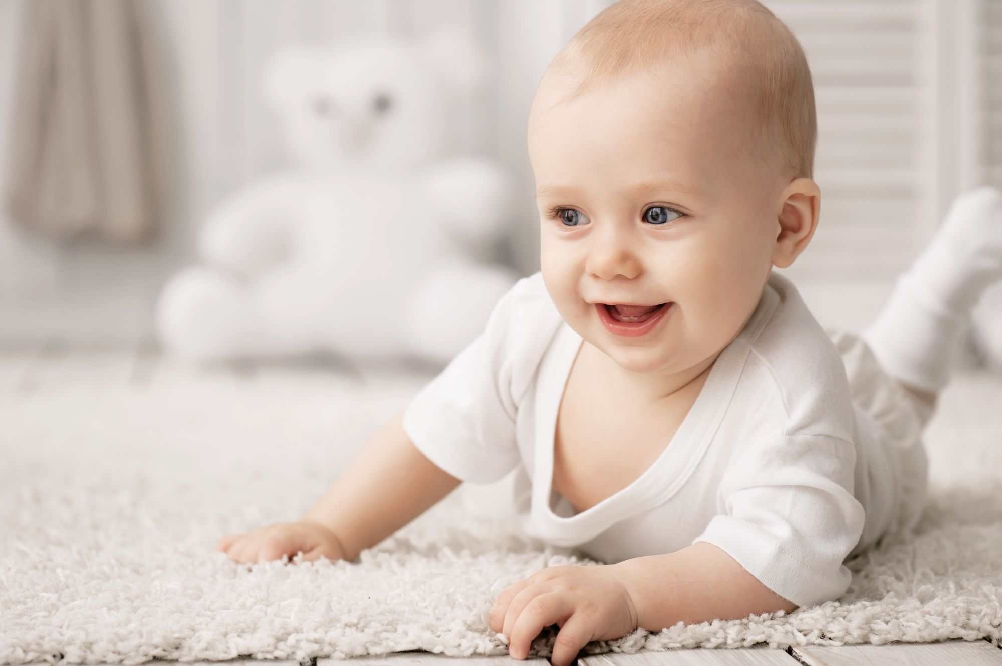 A joyful baby with blue eyes and short hair is crawling on a soft, light-colored rug. The baby is smiling widely, wearing a simple white outfit. In the background, a soft toy bear is faintly visible, adding a warm touch to the scene.