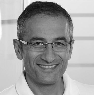 A middle-aged man with short, dark hair and glasses smiles warmly at the camera. He is wearing a white polo shirt, and the background is a light, blurred interior, suggesting a professional setting.