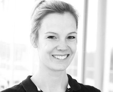 Eine lächelnde Frau mit blonden Haaren und einem schwarzen Oberteil schaut direkt in die Kamera. Der Hintergrund ist hell und unscharf, was den Fokus auf ihr Gesicht legt.