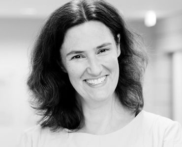 Eine lächelnde Frau mit langen, welligen, dunklen Haaren. Sie trägt ein helles Oberteil und steht in einer hellen Umgebung, die unscharf ist. Ihr freundlicher Gesichtsausdruck vermittelt eine positive Stimmung.