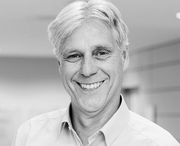 Ein lächelnder Mann mit leicht gewelltem, grauem Haar trägt ein helles Hemd. Der Hintergrund ist unscharf und hell, was auf eine freundliche und einladende Atmosphäre hindeutet.
