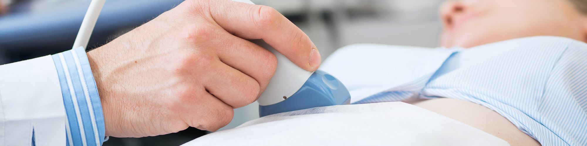 A healthcare professional is using an ultrasound device on a patient's chest, demonstrating a medical examination. The patient is lying down, wearing a blue shirt, while the medical equipment is focused on the area being examined.