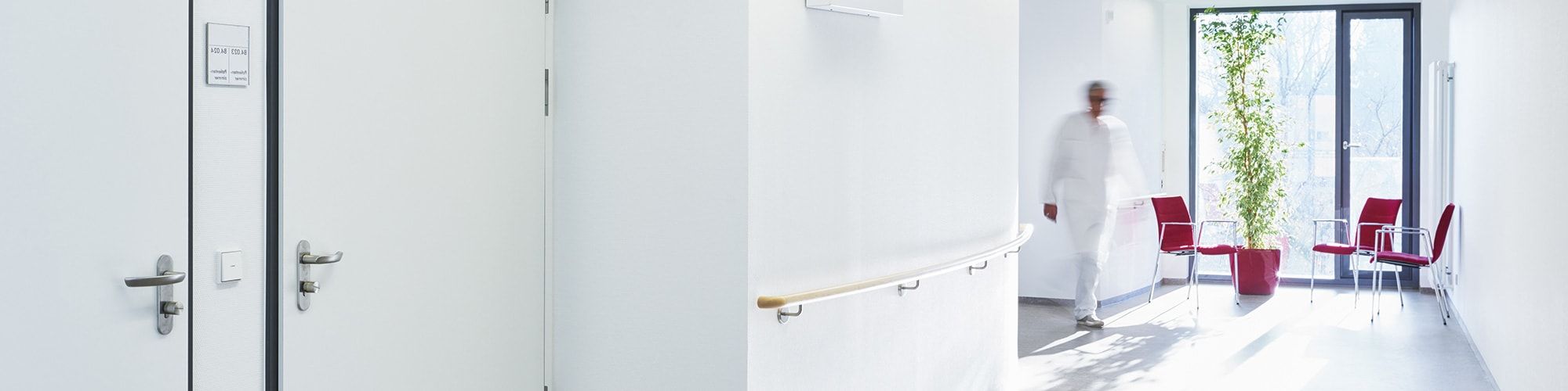 Ein heller Flur in einem medizinischen Umfeld. Zu sehen sind zwei Türen, eine Person in weißem Kittel geht im Hintergrund, und im Vordergrund stehen rote Stühle. Ein Fenster lässt Tageslicht hinein, während eine Pflanze den Raum dekoriert.