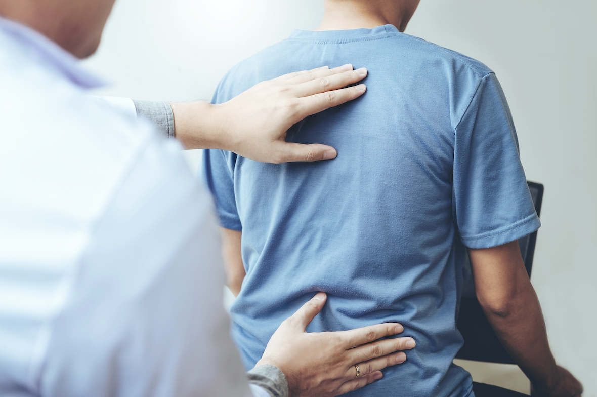 A healthcare professional's hands are positioned on the back of a patient wearing a blue shirt. The scene conveys a therapeutic or supportive interaction, focusing on back treatment or assessment.