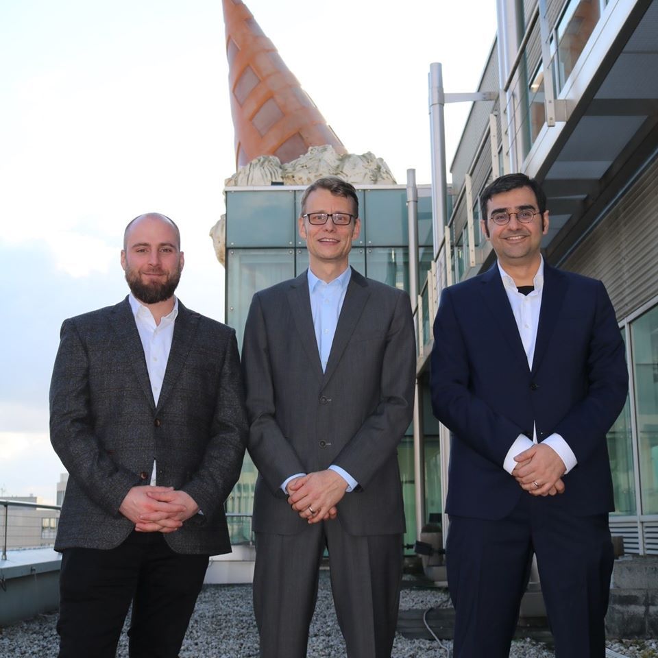 Drei Männer stehen nebeneinander. Der Mann links trägt einen dunklen Anzug mit weißem Hemd, der Mann in der Mitte einen grauen Anzug und der Mann rechts einen dunkelblauen Anzug. Im Hintergrund ist ein Gebäudeteil mit einem auffälligen, kegelförmigen Dach zu sehen.
