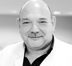A smiling male healthcare professional wearing a white lab coat over a black shirt. He appears confident and welcoming, suggesting a role in patient care or medical leadership. The background is softly blurred, emphasizing his presence.