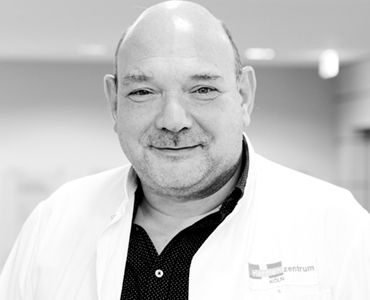 Ein lächelnder Arzt trägt einen weißen Kittel und ein schwarzes Hemd mit Muster. Er steht in einem hellen, modernen medizinischen Umfeld. Der Fokus liegt auf seinem freundlichen Gesichtsausdruck und seiner professionellen Ausstrahlung.