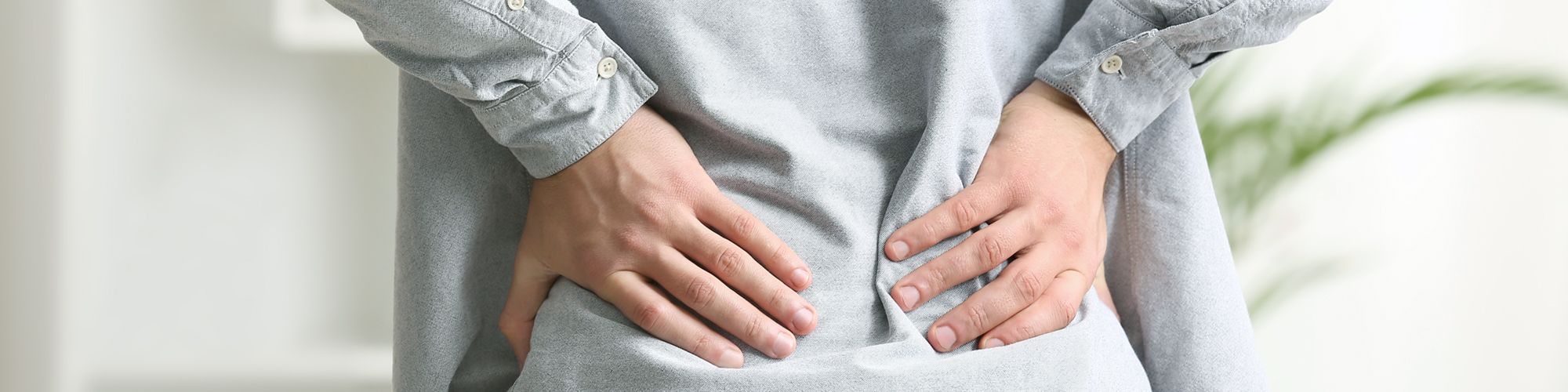 A person is standing with their back to the viewer, wearing a gray shirt. They have both hands placed on their lower back, suggesting discomfort or pain. The background is softly blurred, indicating an indoor setting.