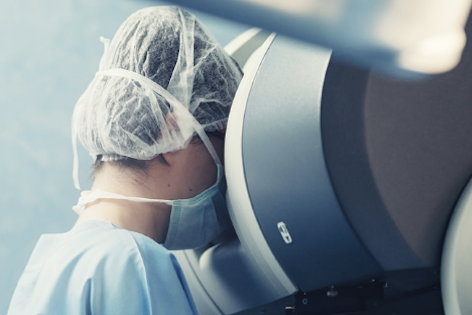 A healthcare professional in surgical attire leans toward a medical machine, focusing intently. They wear a surgical cap and mask, highlighting a clinical setting. The background is softly blurred, emphasizing the professional’s engagement with the equipment.