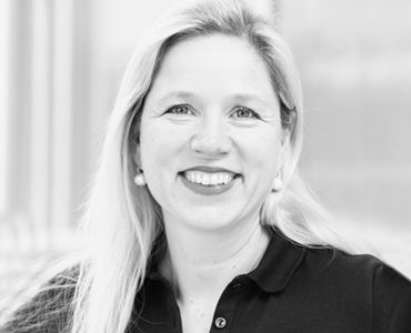 Porträt einer lächelnden Frau mit langen, blonden Haaren, die ein schwarzes Poloshirt trägt. Der Hintergrund ist unscharf, und die Frau schaut direkt in die Kamera.