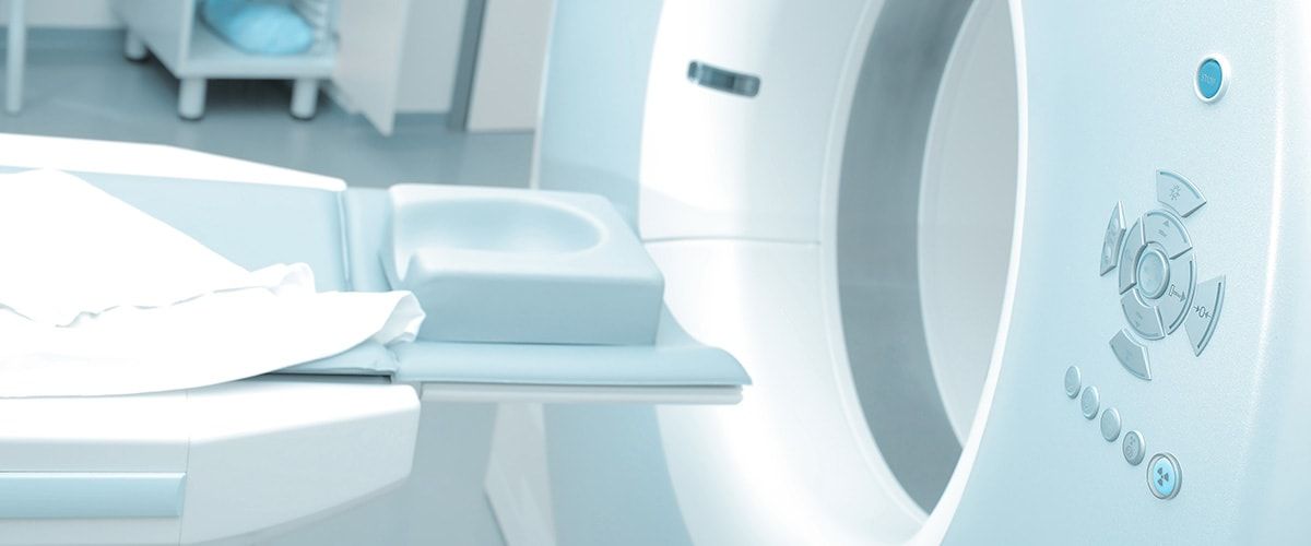 Das Bild zeigt einen Computertomographen (CT-Gerät) in einem klinischen Raum. Es ist eine Liege zu sehen, die in das Gerät führt, sowie Bedienelemente, die zur Steuerung des Geräts verwendet werden. Der Raum hat eine helle und saubere Gestaltung.