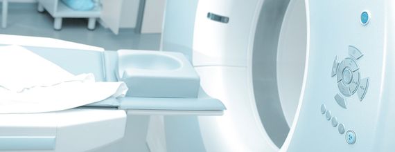 A CT scan machine is shown, featuring a round opening and a flat examination table. The machine has control buttons on the side. A white sheet is placed on the examination table, suggesting preparation for a medical procedure. The space appears clean and clinical.
