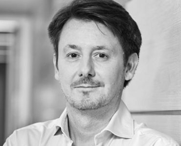 Portrait eines Mannes mit kurzen Haaren und leichtem Bart, der im Vordergrund steht. Er trägt ein Hemd und schaut direkt in die Kamera, mit einer neutralen Miene. Der Hintergrund ist unscharf und vermittelt eine professionelle Atmosphäre.