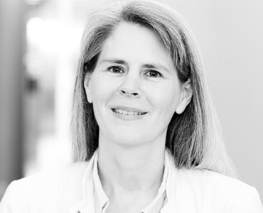 Ein Porträt einer lächelnden Frau mit längeren, hellen Haaren. Sie trägt einen weißen Kittel und hat einen freundlichen Ausdruck. Der Hintergrund ist unscharf und hell, was den Fokus auf ihr Gesicht lenkt.