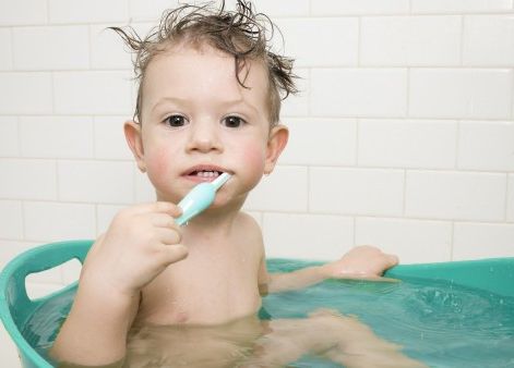 A young child with messy hair sits in a teal tub filled with water, holding a toothbrush and smiling at the camera. The background features white tiled walls, creating a clean bathroom environment.