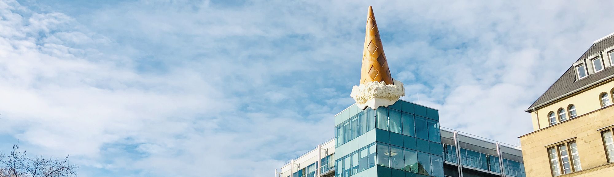 The image shows a modern building topped with a large, whimsical ice cream cone made of a gold-colored cone and white cream. The sky is blue with a few clouds in the background, adding to the playful and striking appearance of this architectural feature.