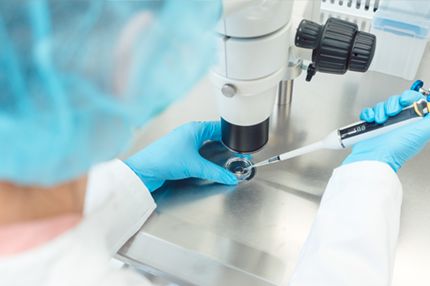 A laboratory technician in a white coat and blue gloves uses a pipette to transfer a sample into a petri dish while observing through a microscope. The setting is a clean lab environment focused on scientific research.