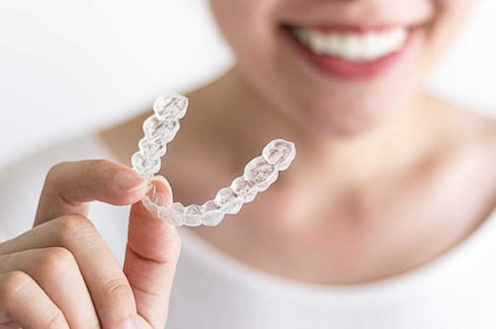 A person is smiling and holding a clear dental aligner in their hand. The aligner is shaped like a mouthguard and is designed for teeth straightening. The background is neutral and focuses on the individual and the aligner.