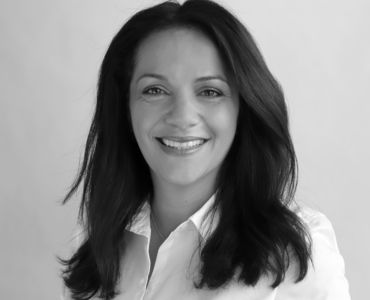 Ein Porträt einer lächelnden Frau mit langen, dunkelbraunen Haaren. Sie trägt ein weißes Hemd und hat eine positive Ausstrahlung. Der Hintergrund ist neutral und hell, was den Fokus auf ihr Gesicht lenkt.