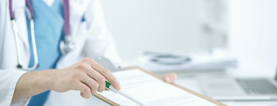 A healthcare professional in a white coat points to a clipboard with documents while sitting at a desk. Medical tools and a computer are partially visible in the background, indicating a clinical setting. The image conveys a focus on patient care and documentation.
