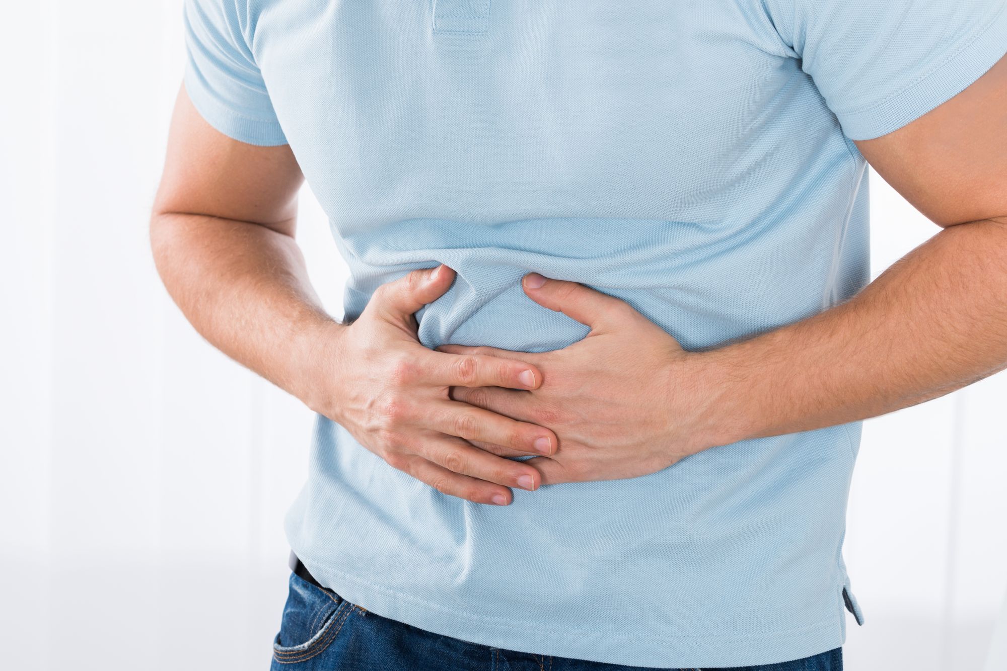 A person in a light blue shirt is standing with their hands on their stomach, appearing to be in discomfort. The setting is simple, with a neutral background. The posture suggests they may be experiencing abdominal pain.
