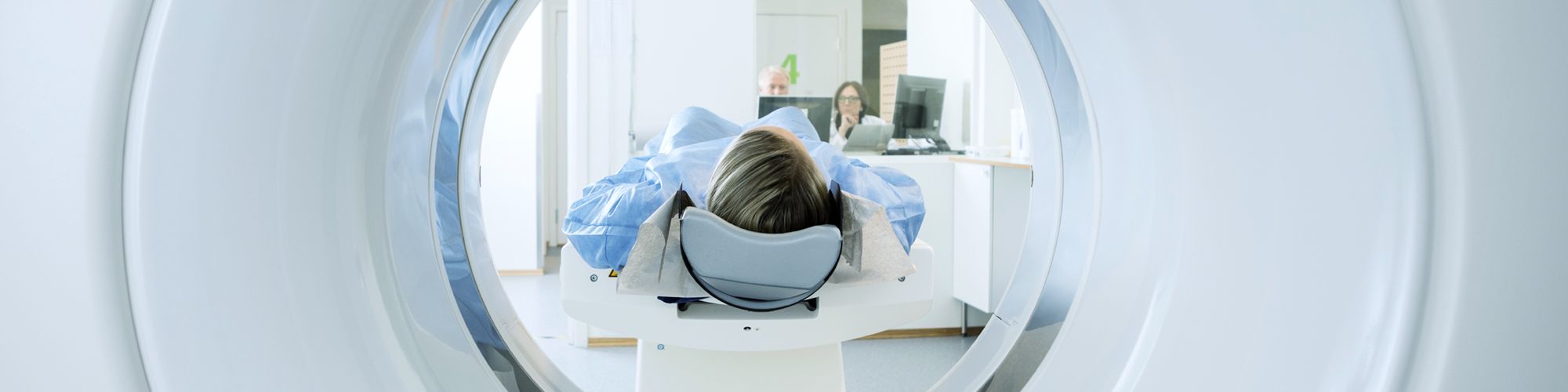 A patient lies on a scanner bed inside a medical imaging machine, partially surrounded by the machine's circular opening. A healthcare professional is visible in the background, preparing for the scan. The environment appears clean and well-lit, typical of a medical facility.