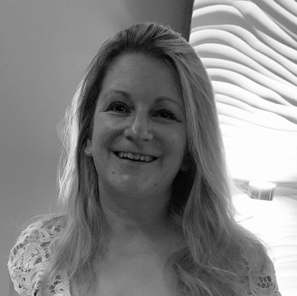 Eine lächelnde Frau mit langen, blonden Haaren steht vor einer strukturierten Wand. Sie trägt ein weißes Oberteil mit Spitzendetails und hat eine freundliche Ausstrahlung.