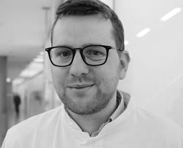 Porträt eines Mannes in einem weißen Kittel und Brille, der freundlich in die Kamera lächelt. Der Hintergrund zeigt eine helle, moderne Umgebung, möglicherweise ein medizinischer Bereich.