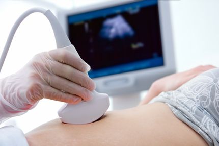 A healthcare professional, wearing gloves, is conducting an ultrasound on a pregnant person's abdomen. An ultrasound machine is visible in the background, displaying a grayscale image. The scene emphasizes prenatal care and the use of ultrasound technology to monitor fetal development.