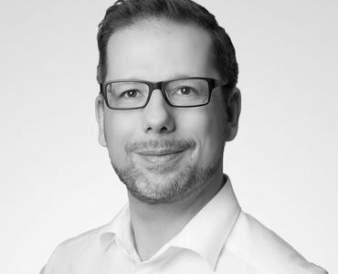 Porträt eines Mannes mit Brille, lächelnd und in einem hellen Oberteil gekleidet. Der Hintergrund ist neutral und hell, was den Fokus auf das Gesicht legt.