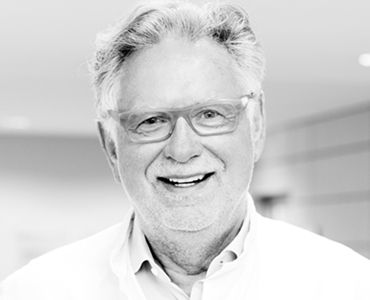 Ein älterer Mann mit grauen Haaren und Brille lächelt freundlich in die Kamera. Er trägt ein weißes Hemd oder eine Arztkittel. Der Hintergrund ist unscharf und hell, was eine positive und freundliche Atmosphäre erzeugt.