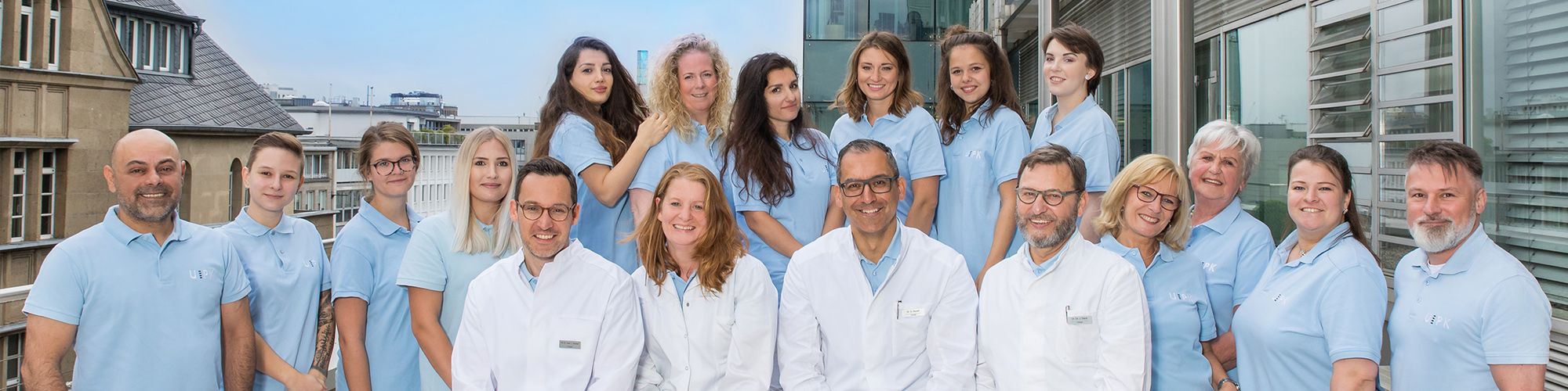 Eine Gruppe von 18 Personen steht vor einem modernen Gebäude. Sie tragen überwiegend hellblaue und weiße Kleidung. Die Gruppe besteht aus Männern und Frauen unterschiedlichen Alters, die freundlich in die Kamera lächeln. Im Hintergrund ist eine städtische Umgebung zu erkennen.