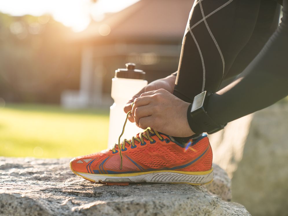 A person is tying the shoelaces of a bright orange running shoe while sitting on a rock. A water bottle is nearby, and the setting appears to be an outdoor area with soft sunlight filtering through trees.