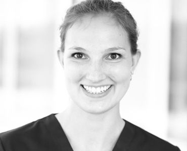 Ein freundlicher, lächelnder Arzt oder Pflegekraft in einem schwarzen Kittel. Sie hat kurze, hellbraune Haare und wirkt freundlich und einladend. Der Hintergrund ist unscharf und hell gehalten.