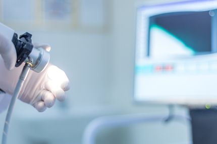 A gloved hand holds a medical instrument emitting light, pointed at a computer screen displaying a graph or waveform. The setting appears to be a healthcare environment, suggesting ongoing medical examination or analysis.