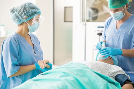 Two medical professionals in scrubs, masks, and gloves are attending to a patient lying on an operating table. One is applying a treatment, while the other observes. The environment appears clinical and sterile, indicating a medical procedure is underway.